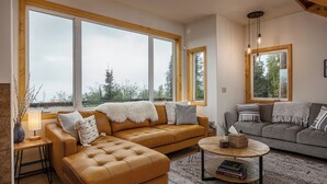A spacious living room with large windows, a brown leather sofa, a grey sofa, and a round coffee table. - Norrsken View - Sauna - Mountains - Deck - Aurora - Family Friendly - Wifi (Fairbanks)
