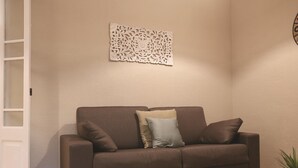 A brown sofa with three cushions against a plain wall, a white door to the left, and a small potted plant to the right. - Sagrada Familia Apartment 14 (Barcelona)