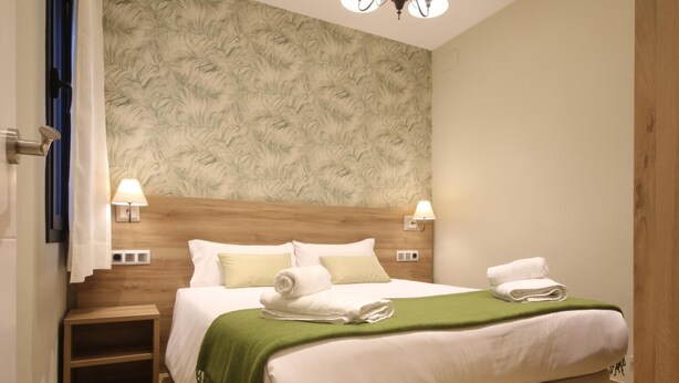 Un lit bien fait, avec du linge de lit blanc et un plaid vert, deux lampes de chevet et une tête de lit en bois.