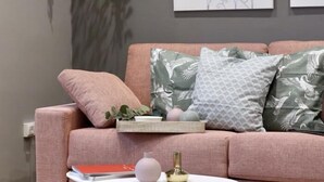 Un salon chaleureux avec un canapé rose, des coussins décoratifs, une petite table sur laquelle se trouvent un vase et un livre, ainsi que des œuvres d’art accrochées au mur.