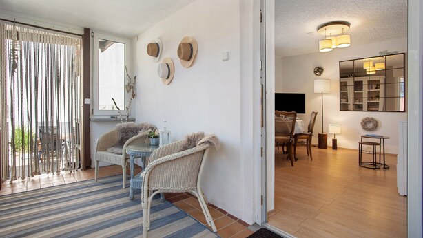 A room with a dining table, chairs, a small side table, a wicker chair, a mirror, and a door leading to another room.