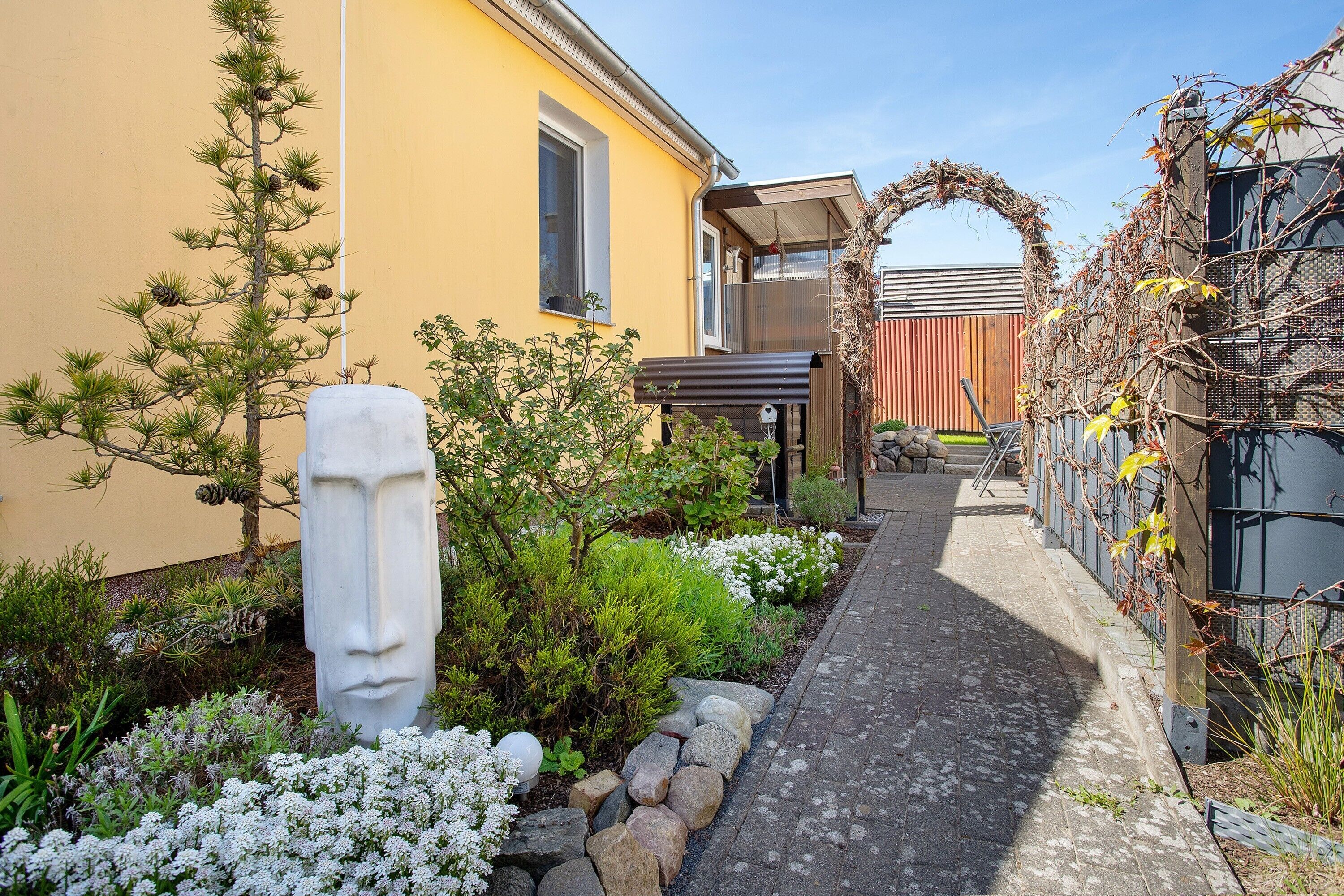 A garden pathway leading to a building with a yellow exterior, a stone face sculpture, and a wooden pergola.