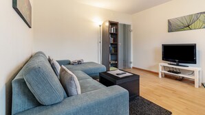 Un salon comprenant un canapé d’angle, un téléviseur à écran plat posé sur un meuble, une bibliothèque et une porte donnant sur une autre pièce.