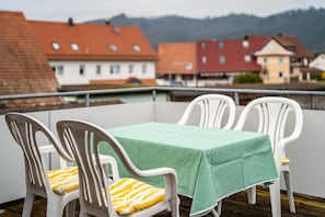 En balkon med et bord og stole, udsigt over en by med bygninger og bjerge i baggrunden.