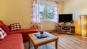 A living room with a red sofa, a coffee table with a remote control, a television on a stand, and a window with curtains. - Apartment 'Kreuzstube' with Mountain View and Wi-Fi (Biberach)