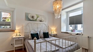 A bedroom with a white bed, a nightstand, a lamp, a window with blinds, and a chandelier. - Apartment 'Ferienwohnung Zur Weinsteige' with Wi-Fi (Ihringen am Kaiserstuhl)
