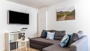 A modern living room with a flat-screen TV, a gray sofa with blue cushions, a small side table with a vase, and a painting of a vineyard on the wall. - Apartment 'Ferienwohnung Zur Weinsteige' with Wi-Fi (Ihringen am Kaiserstuhl)