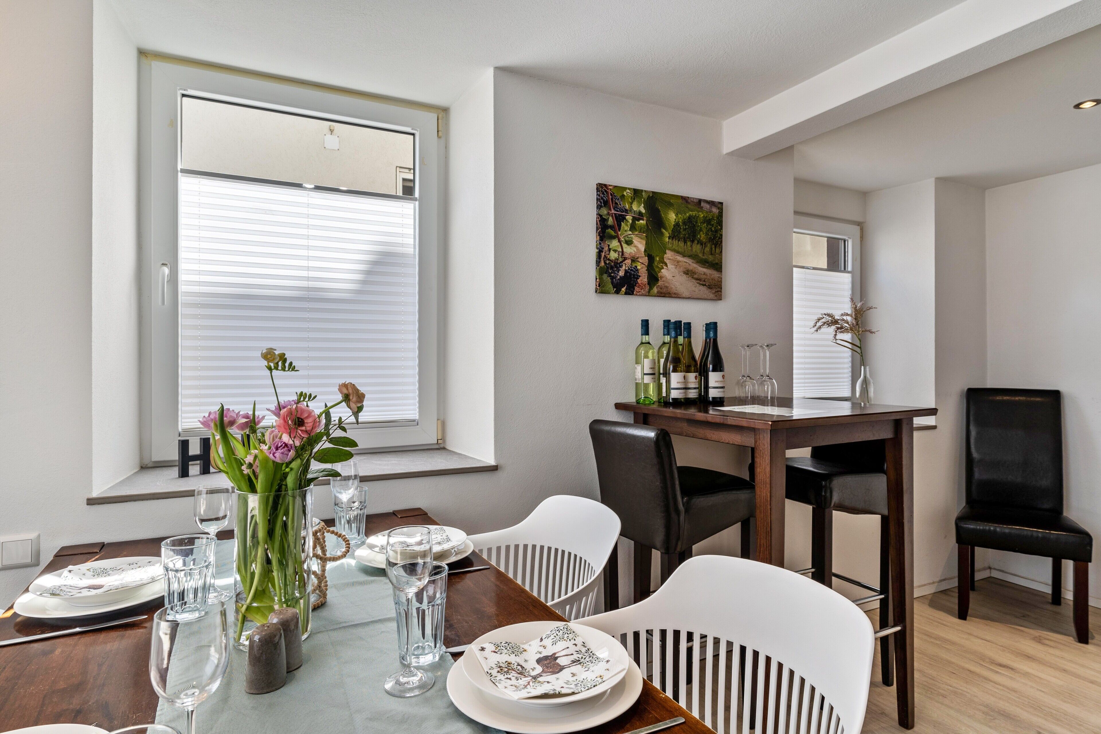 Un comedor con una mesa de madera dispuesta para cuatro personas, sillas blancas, un jarrón con flores y un cuadro en la pared.