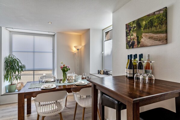 Un comedor con una mesa de madera, sillas blancas, una planta en maceta y una estantería para vinos con botellas y copas.