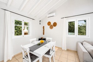 A dining area with a white table, chairs, and a black runner. There are two windows with white curtains, a wall-mounted air conditioner, and a wooden wall decoration.