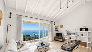 Een moderne woonkamer met een bank, een fauteuil, een salontafel, een tv-meubel en een boekenkast. Er is een grote schuifpui die leidt naar een balkon met uitzicht op zee.