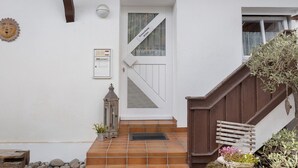 Fachada blanca con una escalera de madera, una puerta con un panel de vidrio diagonal y un farol pequeño en los escalones.