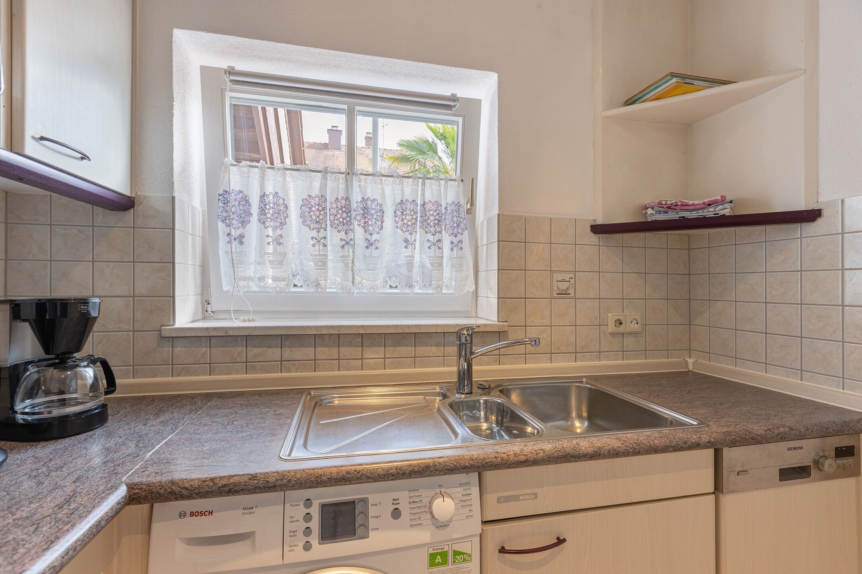 Una cocina con lavavajillas Bosch, una ventana con cortina floral, cafetera y salpicadero de azulejos.