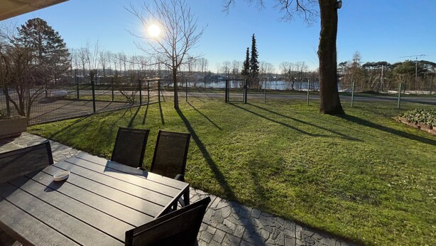 Een terras met een tafel en stoelen, een gazon met een boom en uitzicht op een waterpartij.