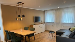 A modern living room with a wooden dining table, green chairs, a black sofa, a TV, and a window with sheer curtains. - Seeterrasse Apartment: Ideal for Cycling, Walks & Relaxation by the Versmolder Seenplatte (Versmold)