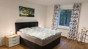 A bedroom with a bed, bedside table, lamp, window with curtains, and a hanging chair. - Seeterrasse Apartment: Ideal for Cycling, Walks & Relaxation by the Versmolder Seenplatte (Versmold)