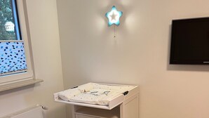 Un cambiador infantil con cajones, una lámpara con forma de estrella, un televisor en la pared y una ventana con persianas.