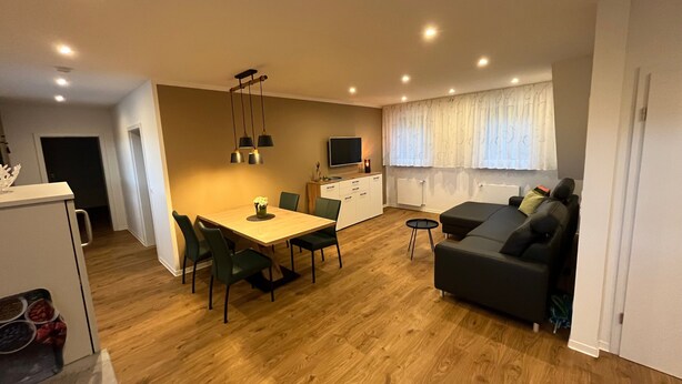 A modern living room with a sofa, dining table, and chairs, a kitchen area with a refrigerator, and a TV. - Seeterrasse Apartment: Ideal for Cycling, Walks & Relaxation by the Versmolder Seenplatte (Versmold)