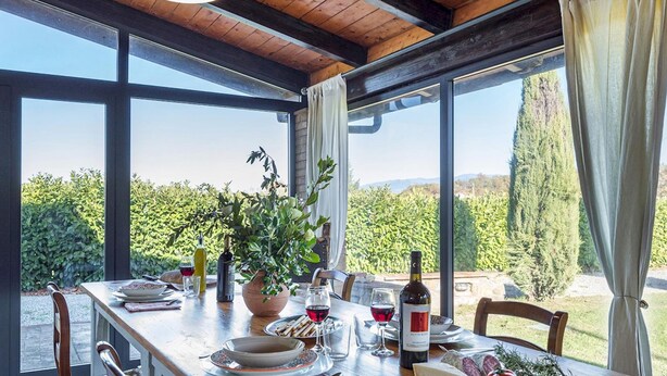 Un coin repas avec une table en bois dressée pour le repas, de grandes fenêtres offrant une vue sur la verdure et les collines, et un luminaire suspendu au-dessus.