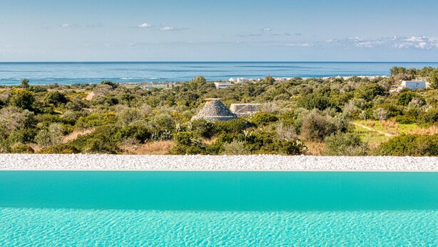 A coastal landscape with a clear turquoise sea, a rocky shoreline, and a dense green area with traditional stone structures. - Villa Porta Azzurra 6 2 Emma Villas (Salve)