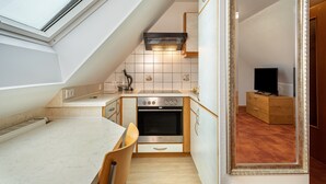 Une cuisine compacte dotée d’un four encastré, un coin repas avec une table et des chaises, et un miroir qui reflète une commode en bois et un téléviseur.