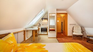 Une pièce de taille modeste comprenant un lit, une chaise, une petite table, un coin cuisine et une porte donnant sur une autre pièce.