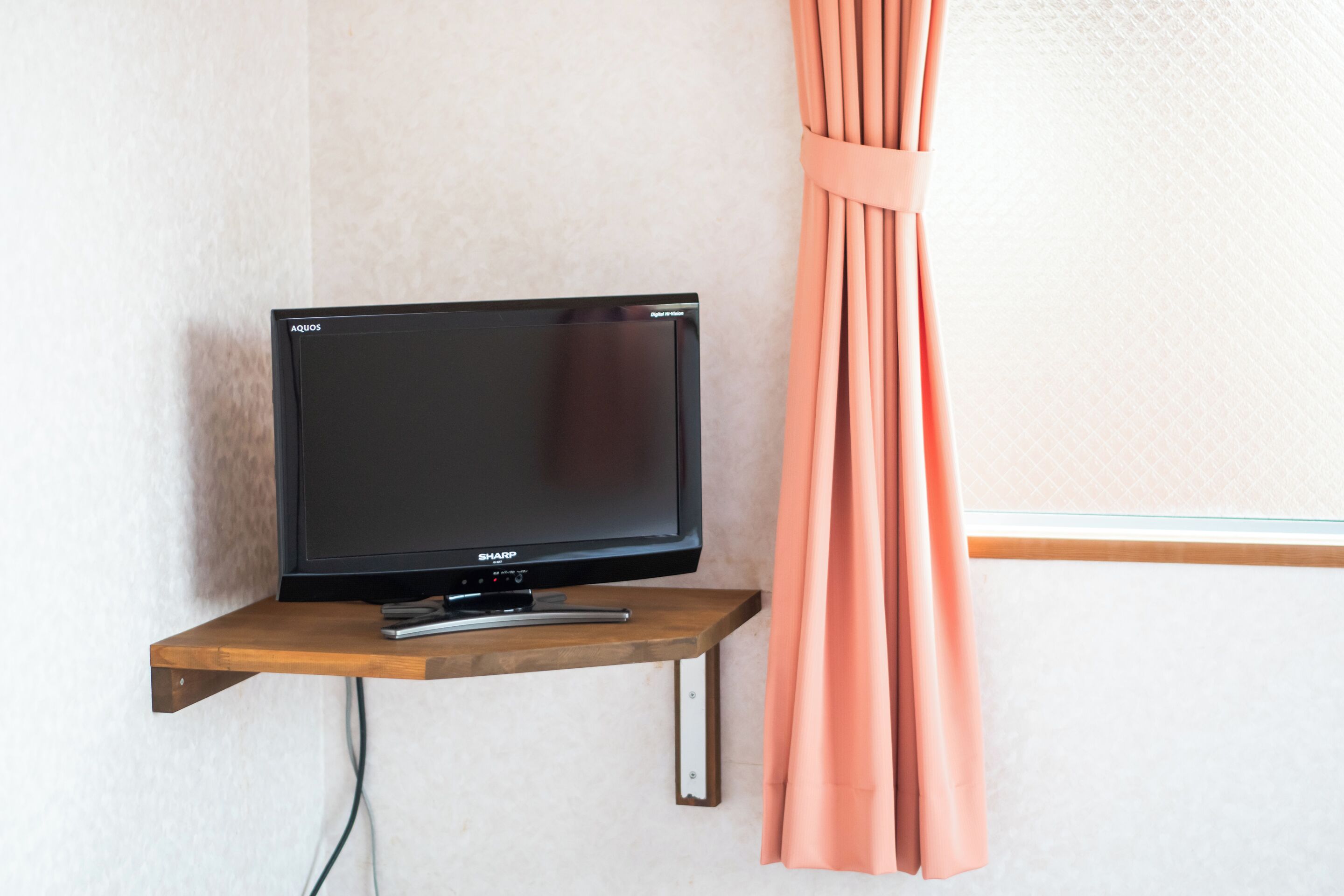 Un téléviseur posé sur une étagère en bois, un rideau et un mur orné d’un motif texturé.