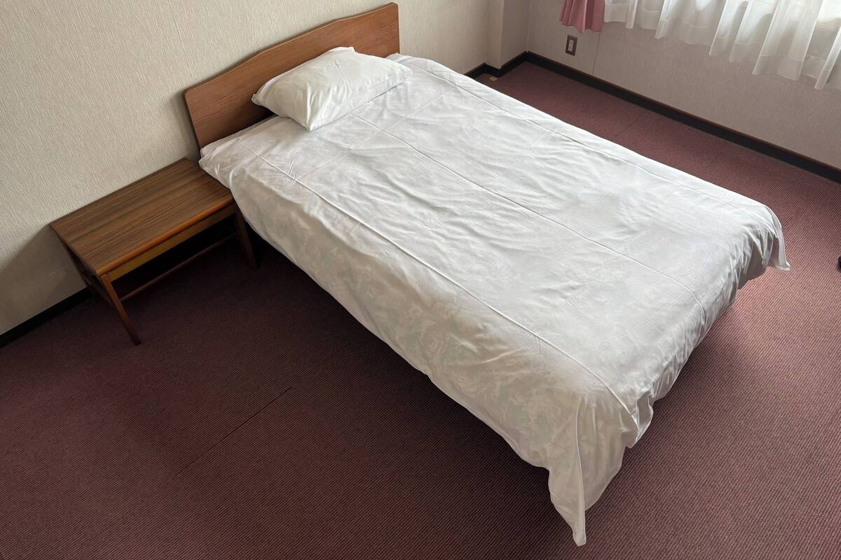 Un lit simple avec une tête de lit en bois, dans une pièce dont les murs sont de couleur unie et dont le sol est recouvert d’une moquette.