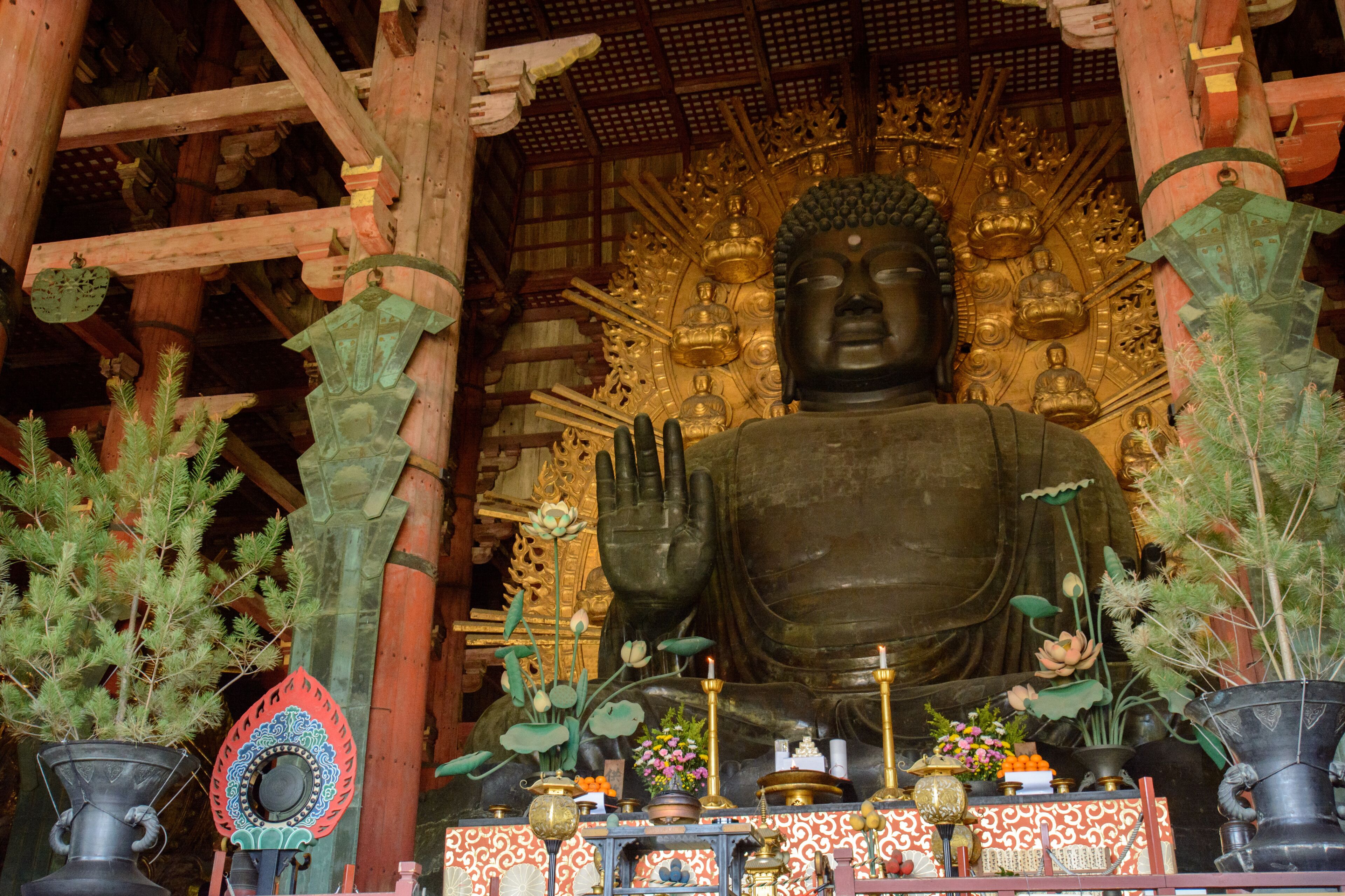 Una gran estatua de Buda en un templo, con vigas de madera y una pared con diseños en el fondo.