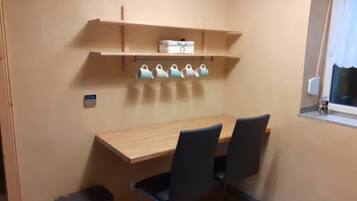 A small room with a wooden table, two chairs, a trash bin, and a shelf with mugs.