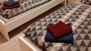 Un petit logement composé d’une seule pièce, avec deux lits, chacun agrémenté de draps et d’oreillers à motifs, une fenêtre avec un rideau et un téléviseur placé au-dessus des lits.