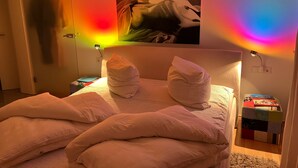 Ein Schlafzimmer mit einem Bett, Kissen, einem Nachttisch, einer Lampe, einem Gemälde und farbigem Licht.
