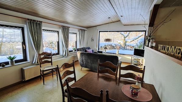 A cozy living room with a wooden table, chairs, a sofa, and a large window with a view of a snowy landscape.