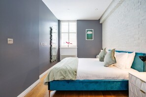 A bedroom with a blue bed, white bedding, a white brick wall, and a window with blinds. - Enchanting Covent Garden Haven (Greater London)