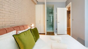Une chambre d’hôtel moderne avec un mur en briques, un lit recouvert de draps blancs, des oreillers verts et blancs, et une porte de douche en verre.