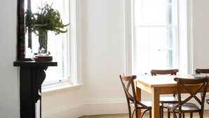 A living room with a dark brown leather sofa, a wooden dining table with four chairs, and a large window with a plant on the sill.