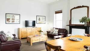 A modern living room with a wooden coffee table, leather sofa, and a dining table with chairs. There are framed pictures on the wall, a flat-screen TV, and a large mirror.
