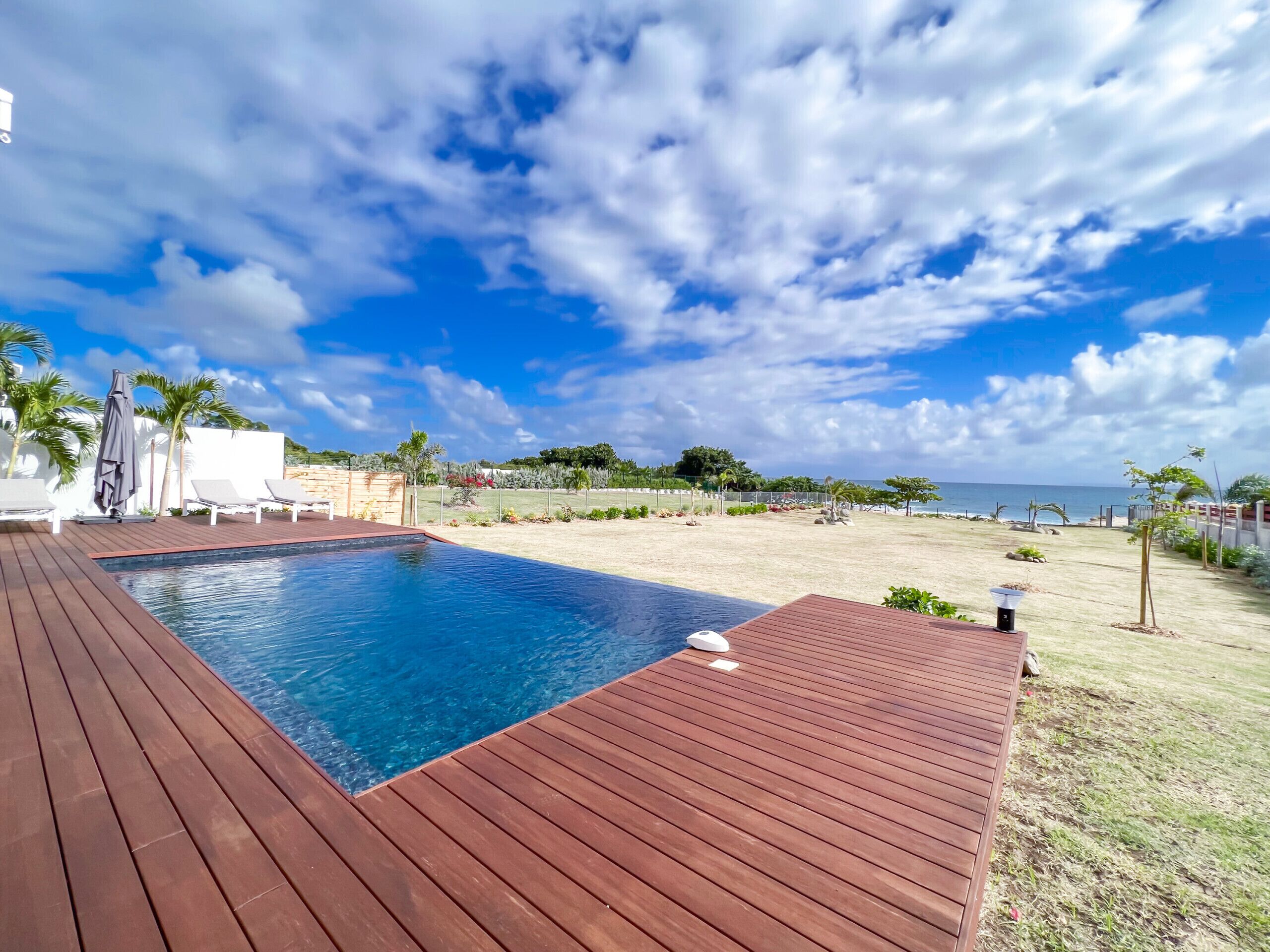 A wooden deck with a pool, lounge chairs, and a view of the beach and ocean.