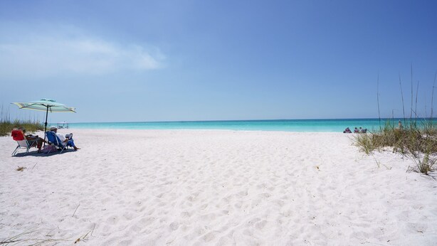 Une plage de sable blanc bordée d’une eau d’un bleu limpide. On aperçoit deux personnes sous un parasol à rayures, et quelques autres au loin.