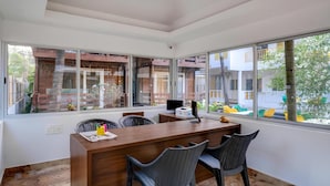 A modern office space with a wooden desk, black chairs, and large windows offering views of a brick building and greenery. - Beach Walk Resort - Goa (Morjim)