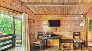 A wooden cabin room with a bed, a desk with a chair, a television, and a window with curtains. - Beach Walk Resort - Goa (Morjim)