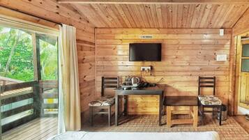 A wooden cabin room with a bed, a desk with a chair, a television, and a window with curtains.