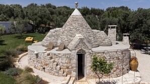 A stone building with a conical roof, a door, and a small garden area with a tree and a yellow bench. - Villa Borghetto Formica (Ostuni)