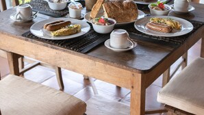 A wooden dining table with various breakfast items, including pastries, bread, and a fruit bowl, set on a veranda with a view of greenery. - Ayawana Private Villa by ARM Hospitality (Tegallalang)