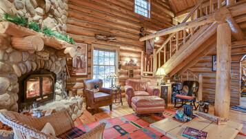A log cabin interior with a stone fireplace, leather furniture, and a wooden staircase.