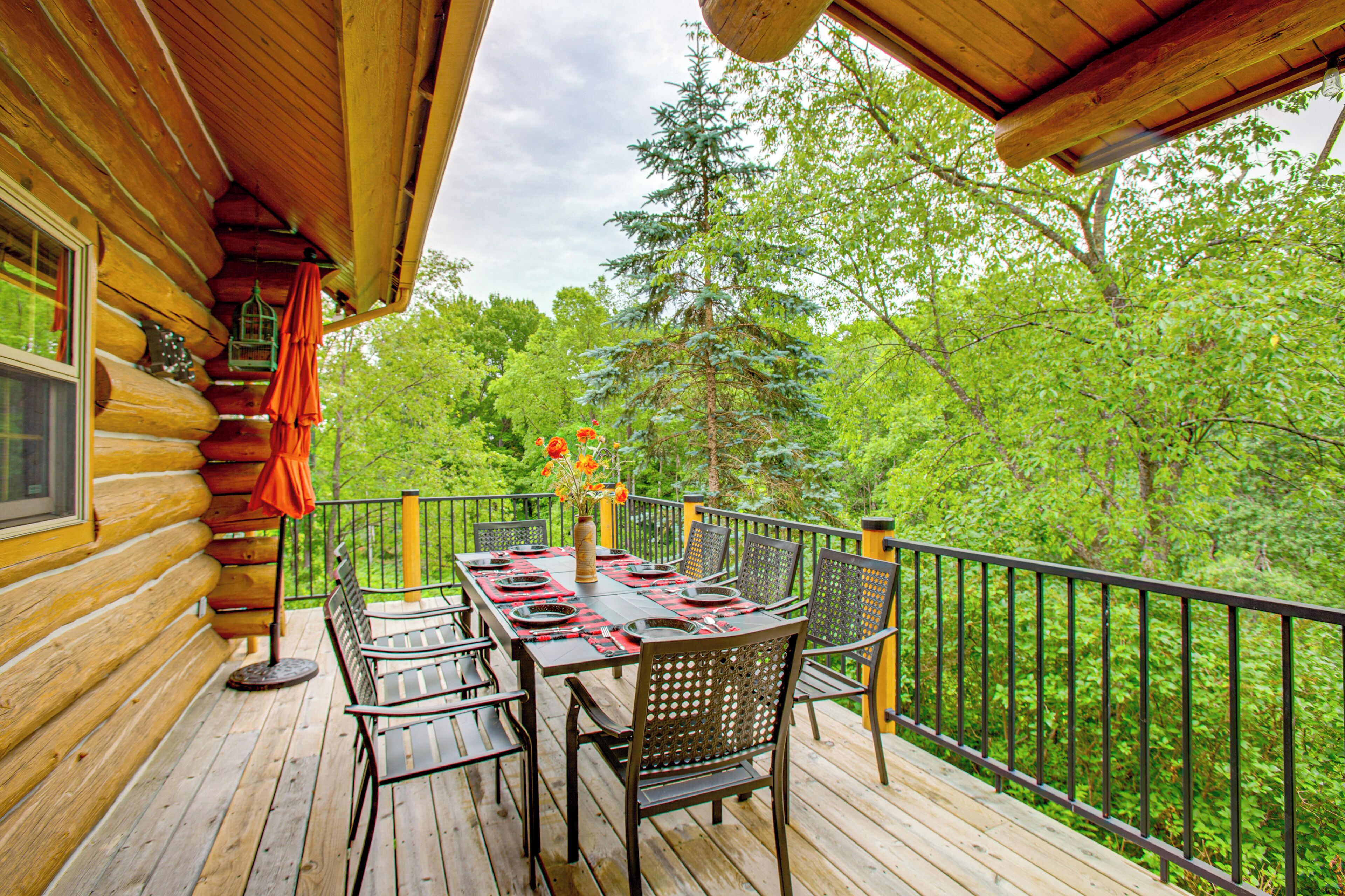 A wooden deck with a table set for dining, surrounded by chairs, overlooking a forested area.