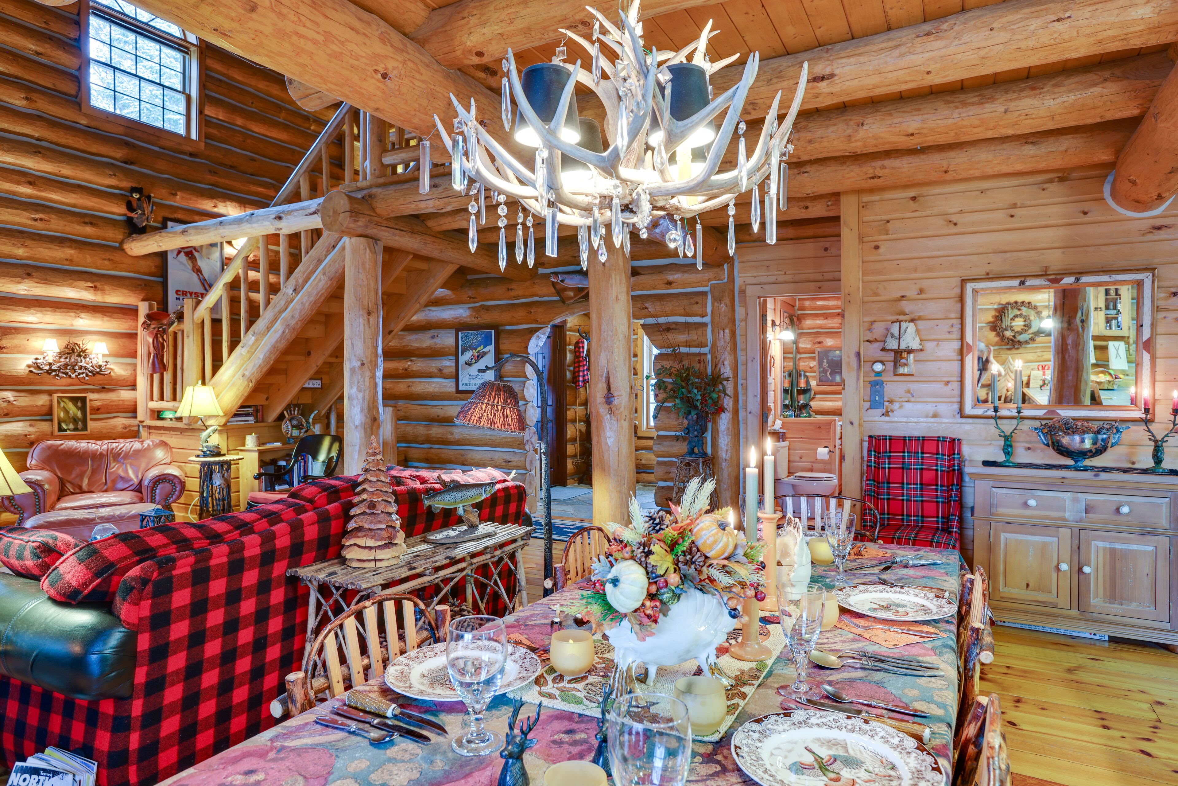 A log cabin interior with a dining table set for a meal, a fireplace, and a staircase leading to an upper level.