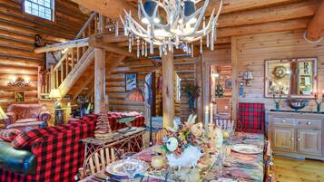 A log cabin interior with a dining table set for a meal, a fireplace, and a staircase leading to an upper level.