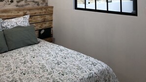 A bedroom with a bed, a wooden bench, a window with a view of a stone wall, and a wall-mounted light fixture.