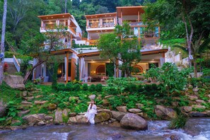 Un hotel de varios pisos con un exuberante jardín y un arroyo en primer plano.